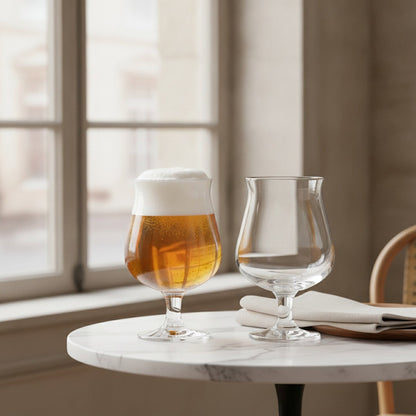 Bulbous stemmed Belgian beer glasses with one filled with golden craft beer near a sunlit window