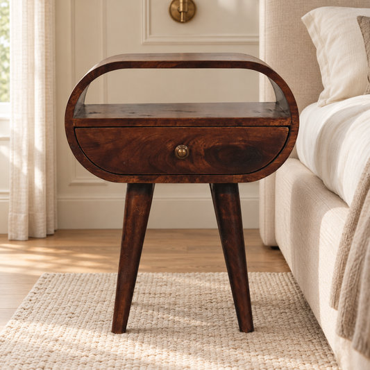 California walnut-finish bedside table with rounded edges, an open shelf and brass drawer knob beside an ivory bed.